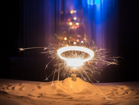 Glittering burning sparkler on snow with blurred Christmas tree on dark background. New Year Holiday concept with empty space for your text. Selective focusの写真素材