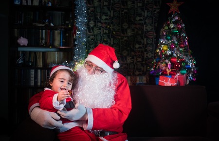 Merry Christmas and happy holidays! Cute little child girl and Santa Claus sitting near glowing fir tree. Holiday conceptの写真素材