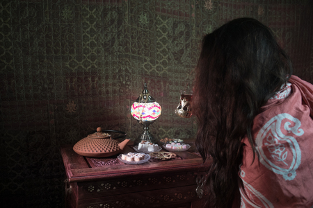 Glass of black tea in woman`s hand at Low light lounge interior with carpet. Eastern tea concept. Eastern snacks on vintage wooden surface. Selective focusの写真素材