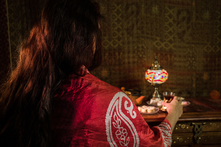 Glass of black tea in woman`s hand at Low light lounge interior with carpet. Eastern tea concept. Eastern snacks on vintage wooden surface. Selective focusの写真素材