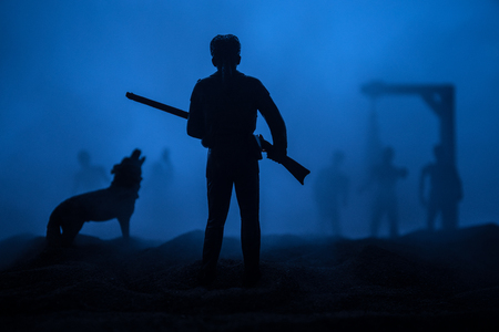 Man with riffle against zombie attack. Zombie apocalypse. Scary view of blurred zombies at cemetery and spooky cloudy sky with fog. Horror Halloween concept.の写真素材