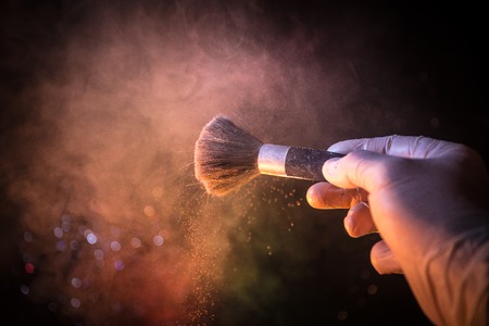 Makeup brush in hand with cosmetic powder on dark background with light and smoke. Powder splash on...