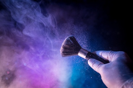 Makeup brush in hand with cosmetic powder on dark background with light and smoke. Powder splash on...