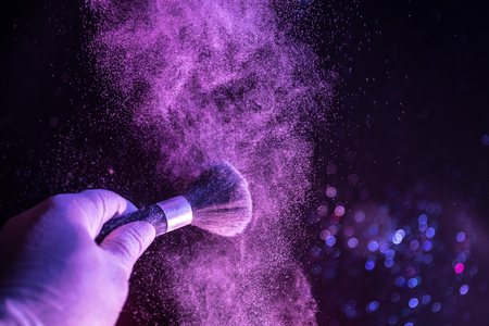 Makeup brush in hand with cosmetic powder on dark background with light and smoke. Powder splash on dark. Selective focusの写真素材