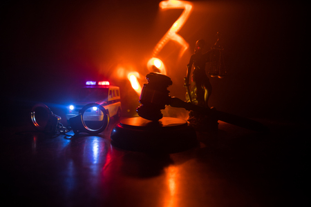 Law theme, mallet of the judge on wooden desk with Lady Justice Statue. Silhouette of handcuffs with police car on backside. Law gavel on dark foggy background with light.の写真素材