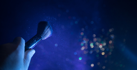Makeup brush in hand with cosmetic powder on dark background with light and smoke. Powder splash on...