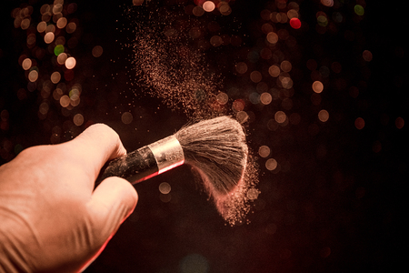 Makeup brush in hand with cosmetic powder on dark background with light and smoke. Powder splash on...