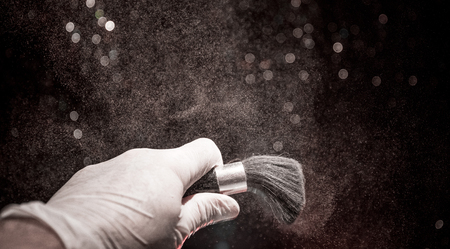 Makeup brush in hand with cosmetic powder on dark background with light and smoke. Powder splash on dark. Selective focusの写真素材