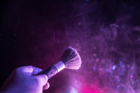 Makeup brush in hand with cosmetic powder on dark background with light and smoke. Powder splash on dark. Selective focusの写真素材