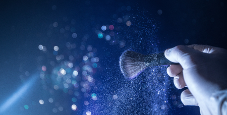 Makeup brush in hand with cosmetic powder on dark background with light and smoke. Powder splash on...