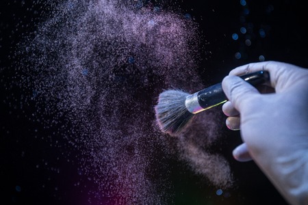 Makeup brush in hand with cosmetic powder on dark background with light and smoke. Powder splash on...