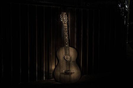 An wooden acoustic guitar is against a grunge textured wall. The room is dark with a spotlight for...