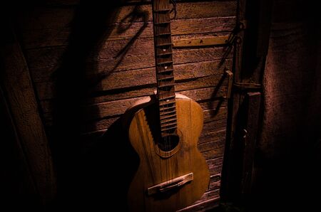 An wooden acoustic guitar is against a grunge textured wall. The room is dark with a spotlight for your copyspace. old broken guitar