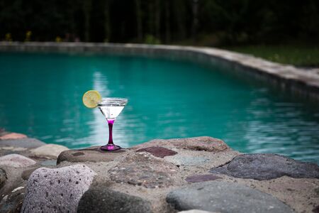 Glass of martini cocktail at swimming pool with forest trees on background. Selective focus. Season and holidays concept.の写真素材