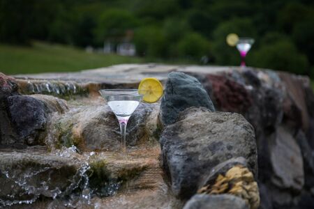 Glass of martini cocktail at mini waterfall with round stones. Selective focus. Season and holidays...