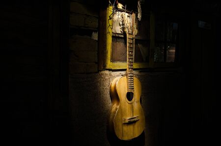An wooden acoustic guitar is against a grunge textured wall. The room is dark with a spotlight for your copyspace. old broken guitarの写真素材