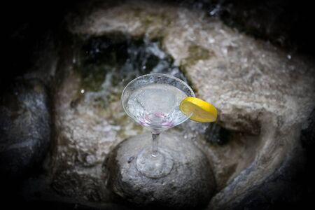 Glass of martini cocktail at mini waterfall with round stones. Selective focus. Season and holidays concept.の写真素材