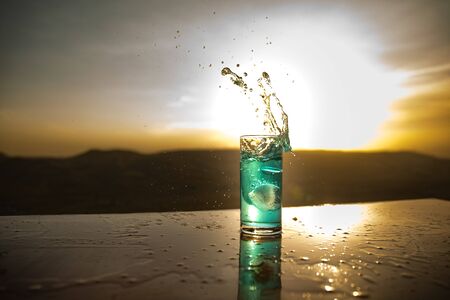 Cooling summer drink concept. Cocktail glasses splashing on sunset. Close up wet glass of cold drink...