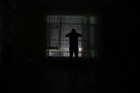 Silhouette of a man standing at a window inside the room. Dark mood conceptual imageの写真素材