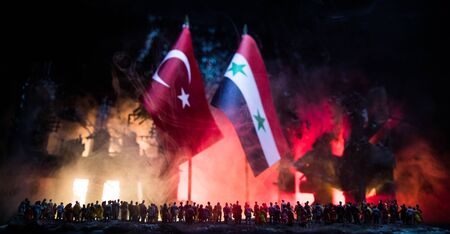Closeup, flags of Syria and Turkey against destroyed city at night. Decorated diorama. Crowd looking...