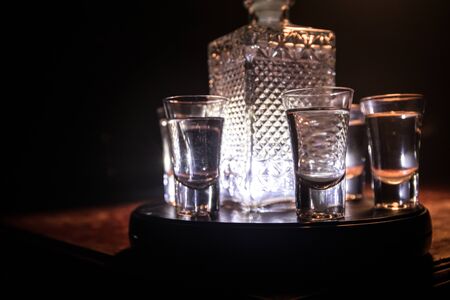A beautiful carafe with vodka stands on a wooden table with glasses. Dramatic toned foggy background with light. Selective focus. Club drink concept.の写真素材