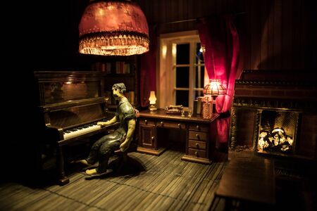 A realistic dollhouse living room with furniture and window at night. Man sitting at the old piano in dark room. Selective focus.の写真素材
