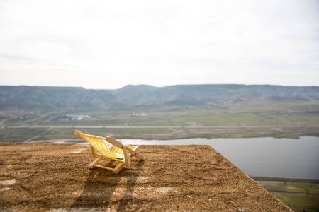 Miniature doll lounger on sand at balcony with view on mountains and lake. Corona virus stay home concept. Funny decoration. Stay at home to reduce risk of infection. Selective focus.の写真素材