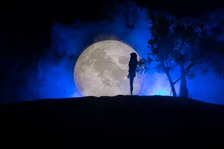 Girl standing alone behind big full moon. Dark toned foggy background. Loneliness bad mood conceptの写真素材