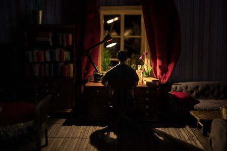 A realistic dollhouse living room with furniture and window at night. Man sitting on table in dark...