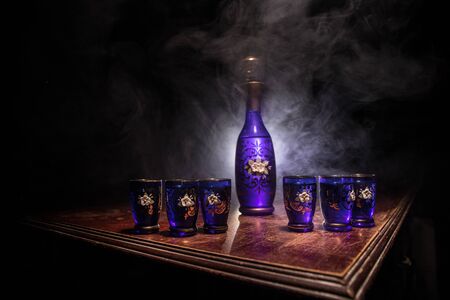 A beautiful blue carafe with vodka stands on a wooden table with glasses. Dramatic toned foggy...