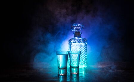 A beautiful carafe with vodka stands on a wooden table with glasses. Dramatic toned foggy background with light. Selective focus. Club drink concept.の写真素材