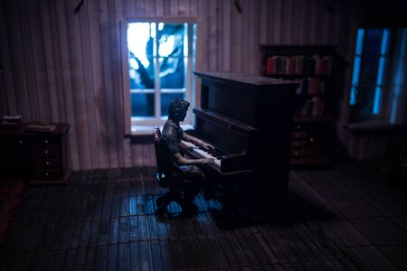 A realistic dollhouse living room with furniture and window at night. Man sitting at the old piano in dark room. Selective focus.の写真素材