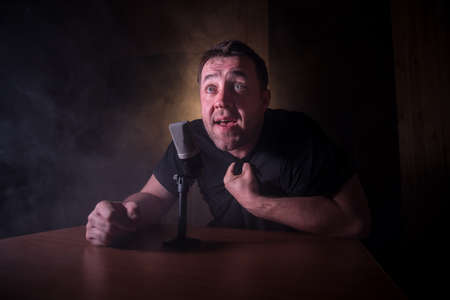 Caucasian man news broadcaster sat at a desk with microphone in dark room. Performer with microphone. Male musician singing into a microphone. Shot on a black background. Dark atmosphereの写真素材