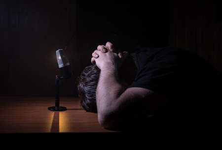 Caucasian man news broadcaster sat at a desk with microphone in dark room. Performer with microphone. Male musician singing into a microphone. Shot on a black background. Dark atmosphereの写真素材