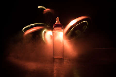 Close up of baby bottle at wooden table on dark toned foggy background with light. Creative artwork decorated photo. Selective focus.の写真素材