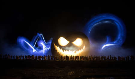 Halloween concept. Blurred silhouette of giant Jack-o-lantern pumpkin with scary smiling face behind crowd at night. People looks at a big pumpkin at night. Selective focus.の写真素材