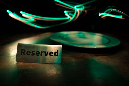 Reserved sign on the table. A tag of reservation placed on the wood table. Metal tag with reservation on dark. Reserved table in a restaurant. Creative concept with colorful lights. Selective focusの写真素材