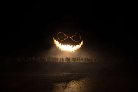 Halloween concept. Blurred silhouette of giant Jack-o-lantern pumpkin with scary smiling face behind crowd at night. People looks at a big pumpkin at night. Selective focus.の写真素材
