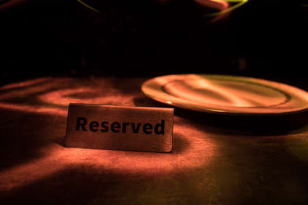 Reserved sign on the table. A tag of reservation placed on the wood table. Metal tag with reservation on dark. Reserved table in a restaurant. Creative concept with colorful lights. Selective focusの写真素材