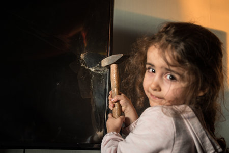 Cute little girl standing in front of a TV with broken screen holding a hammer. Home insurance concept. Child smashed led TV screen with a hammerの写真素材