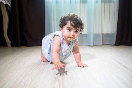 Baby Girl In Summer Dress Sit on wooden floor inside room. Baby girl sitting near window