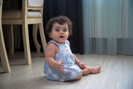 Baby Girl In Summer Dress Sit on wooden floor inside room. Baby girl sitting near window