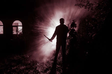 Silhouette of killer with knife standing in the dark forest with light. Horror halloween concept. strange silhouette in a dark spooky forest at nightの写真素材