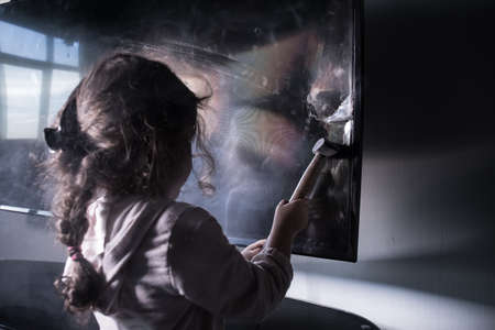 Cute little girl standing in front of a TV with broken screen holding a hammer. Home insurance concept. Child smashed led TV screen with a hammerの写真素材