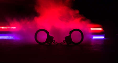Police raid at night and you are under arrest concept. Silhouette of handcuffs with police car on backside. Image with the flashing red and blue police lights at foggy background. Selective focusの写真素材