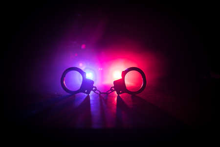 Police raid at night and you are under arrest concept. Silhouette of handcuffs with police car on backside. Image with the flashing red and blue police lights at foggy background. Selective focusの写真素材