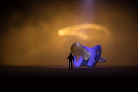 Creative concept. Man silhouette standing at giant baby pacifier on wooden table with low key toned background. Selective focusの写真素材