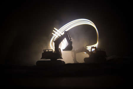Construction site on a city street. A yellow digger excavator parked during the night on a construction site. Industrial concept table decoration on dark foggy toned background. Selective focusの写真素材