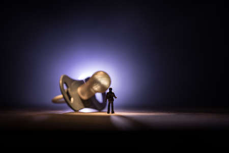 Creative concept. Man silhouette standing at giant baby pacifier on wooden table with low key toned background. Selective focusの写真素材