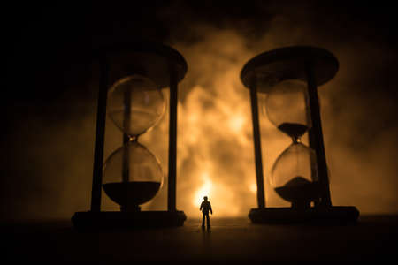 Time concept. Silhouette of a man standing between hourglasses with smoke and lights on a dark...